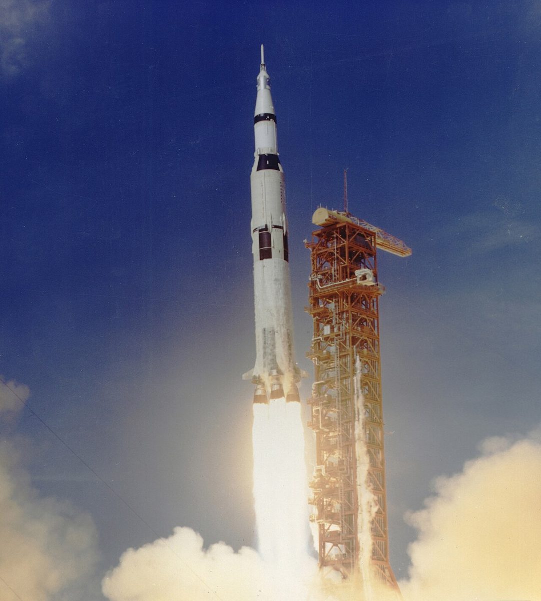 The Saturn V rocket carrying Apollo 11 climbs past its launch tower on a column of flame.