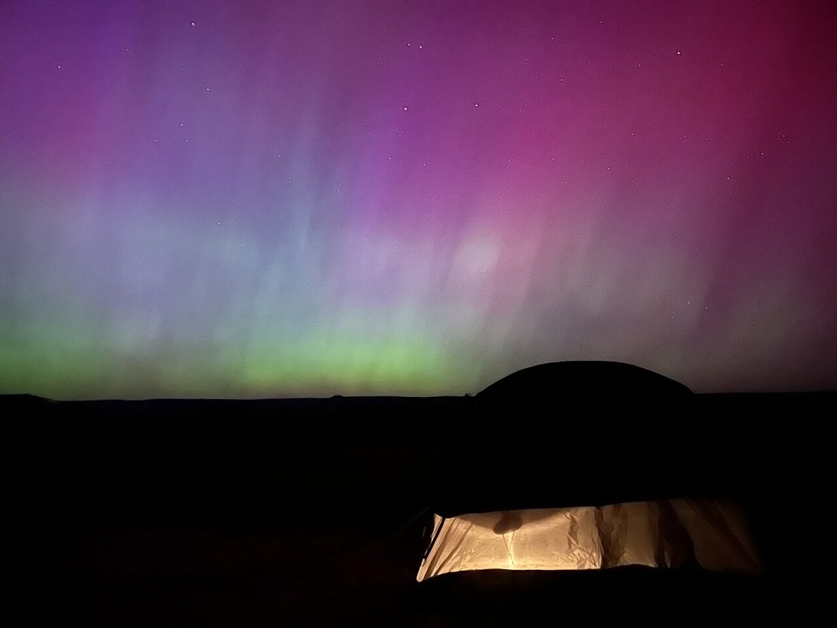 Pink, green and blue northern lights glowing above a dark desert horizon and a lit tent.