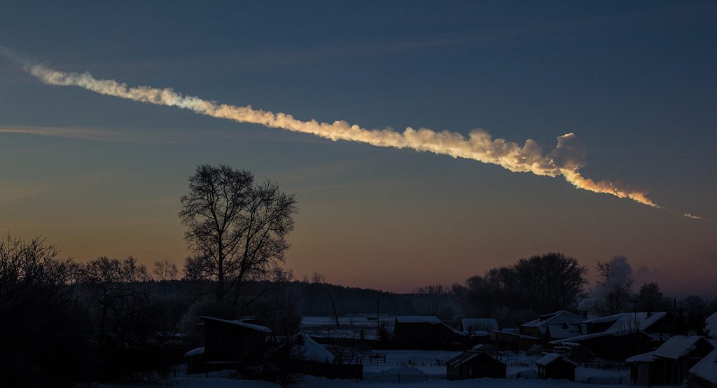The smoky trail left by the Chelyabinsk meteor stretches across a pale morning sky above snowy rooftops and trees.