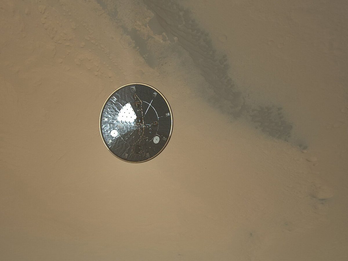 Curiosity's round heat shield falling away toward the tan surface of Mars, seen from the descending rover.