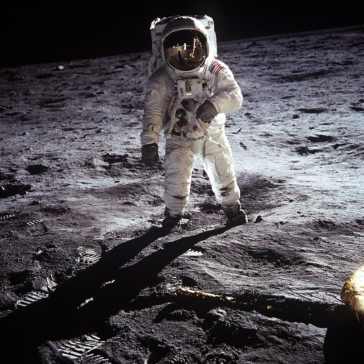 Buzz Aldrin stands on the Moon in his spacesuit, with Neil Armstrong reflected in his visor.