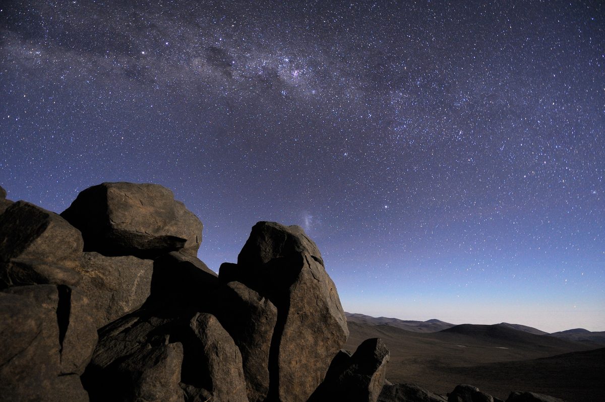 The Milky Way arches over boulders and dark hills in the Atacama Desert at twilight.