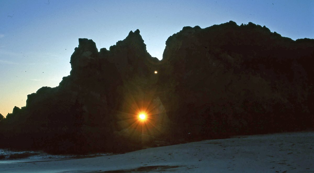 The setting Sun shines through the hole in Keyhole Arch at Pfeiffer Beach, the rock a dark silhouette against the dusk sky.