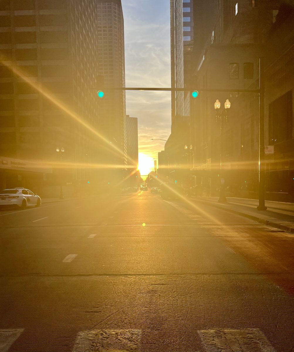 The Sun sets straight down a Chicago Loop street between tall buildings, flooding the road with golden light.