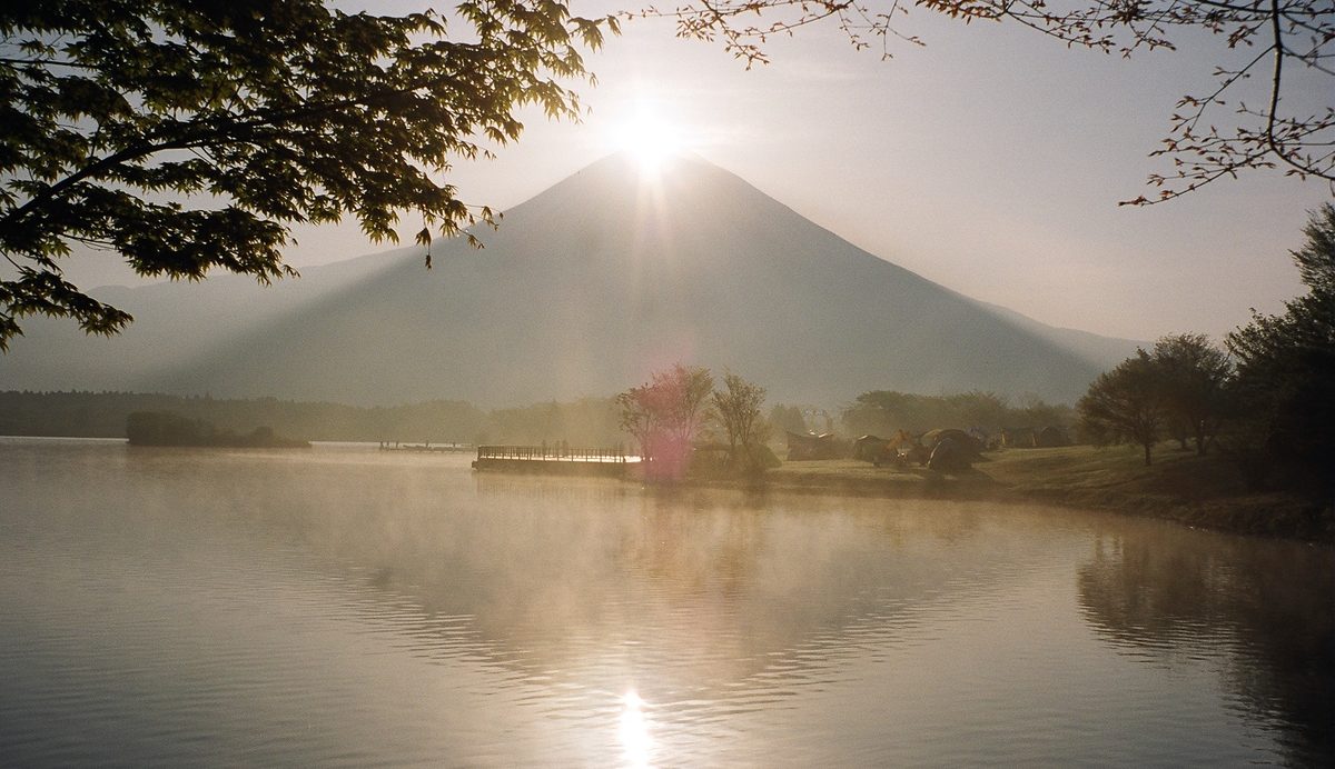 The rising Sun sits exactly on the summit of Mount Fuji above misty Lake Tanuki, its light reflected in the still water.