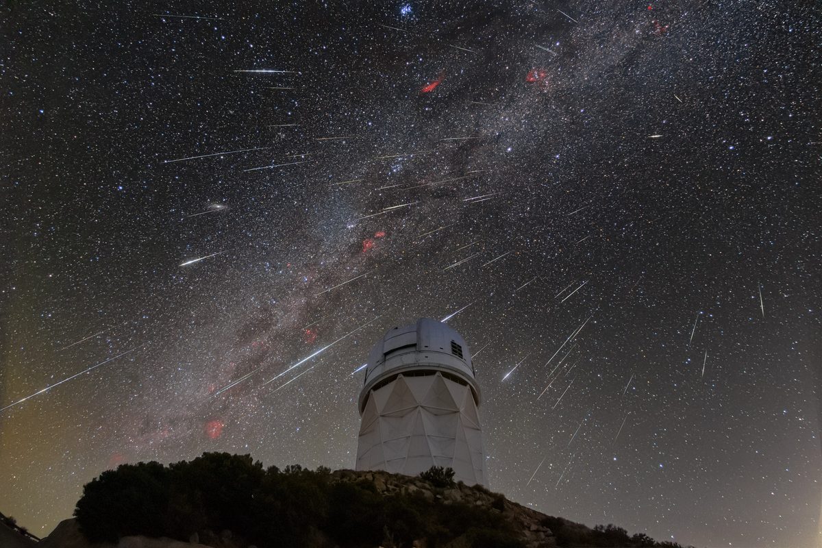 Dozens of Geminid meteors streak across the Milky Way above a white telescope dome on a desert mountaintop.