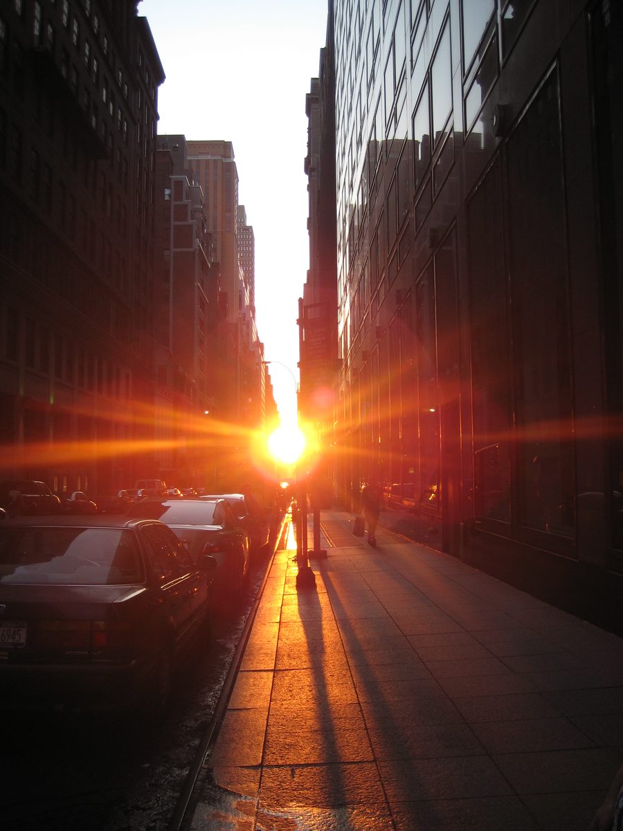The setting Sun blazes at the end of a Manhattan cross street, lighting the sidewalk between tall buildings.