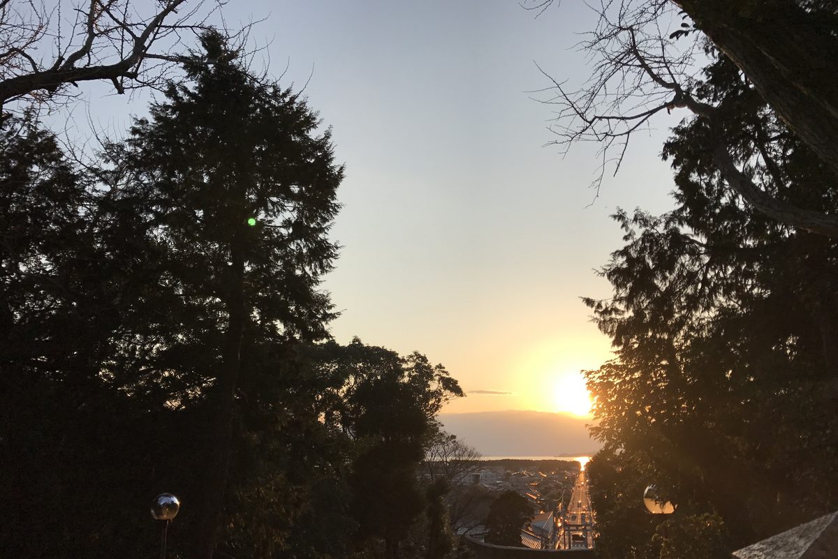 The low Sun shines down the long straight approach road below Miyajidake Shrine toward the sea, lighting the road gold.