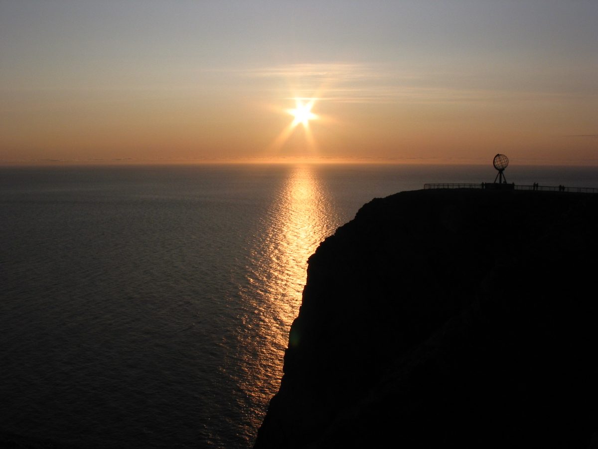 The midnight Sun hangs above the Arctic Ocean beside the dark cliff of North Cape and its globe monument.