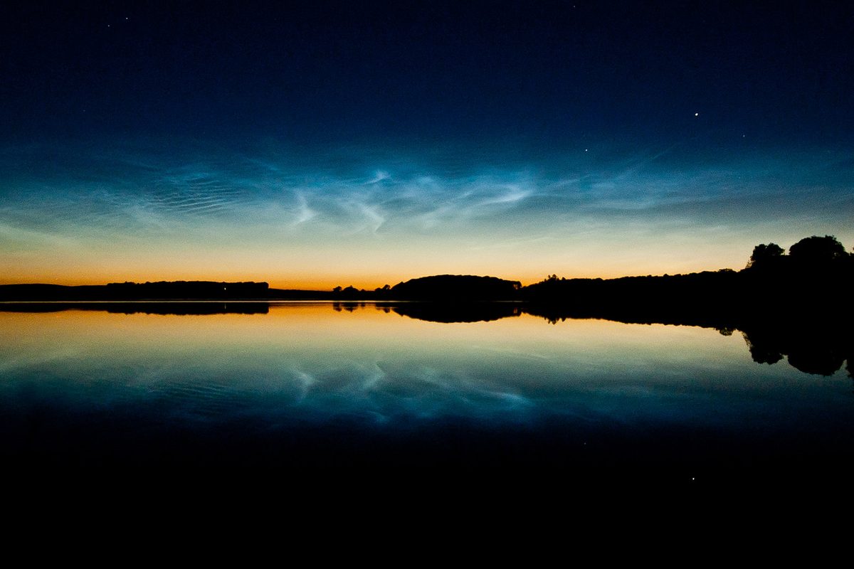 Electric blue rippling noctilucent clouds glow above the twilight horizon, mirrored in the still water of a Scottish loch.