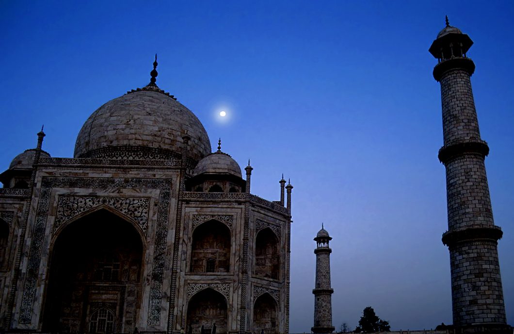 A nearly full Moon shines in the deep blue evening sky between the marble dome and a minaret of the Taj Mahal.