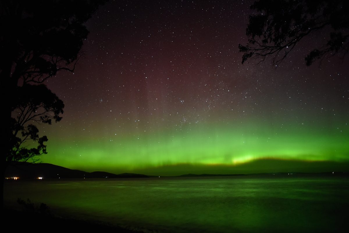 A green and red aurora australis glows low over the water on a starry night in southern Tasmania.