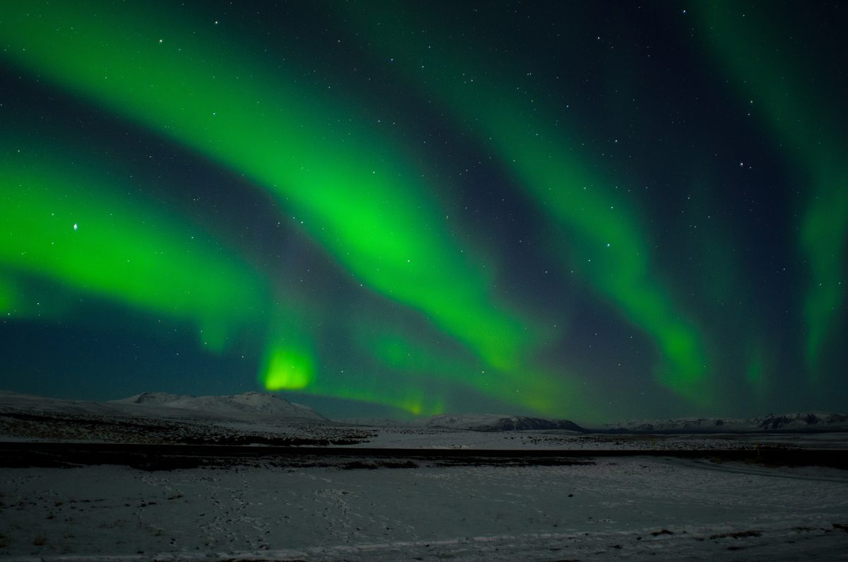 Bright green bands of the northern lights stretch across a starry sky over snowy ground in Þingvellir National Park.