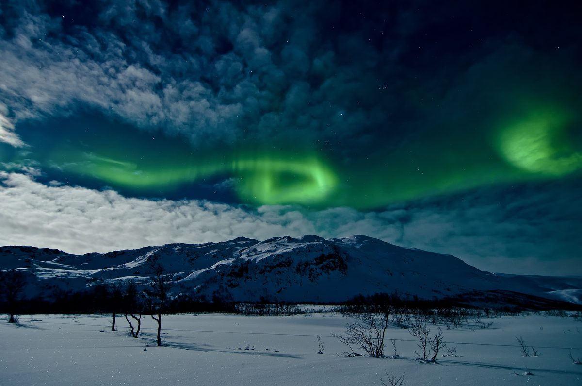 Green rings of the northern lights swirl through moonlit clouds above snowy mountains near Tromsø.
