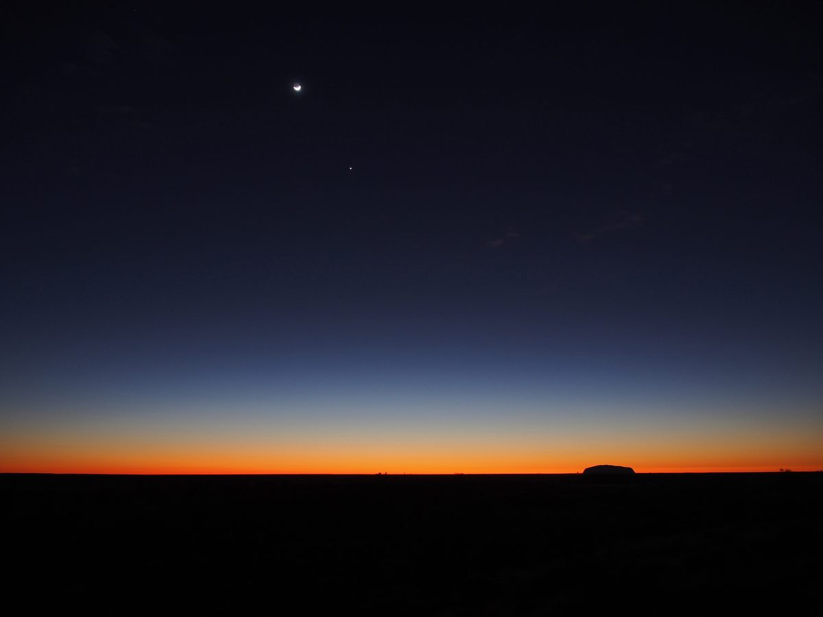 A crescent Moon and a bright planet shine in the deep blue twilight above the dark outline of Uluru on the horizon.