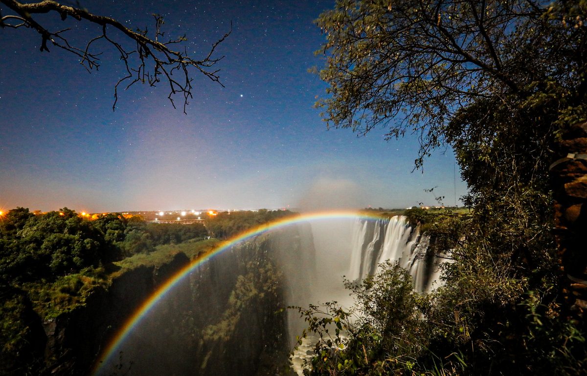A lunar rainbow arches across the moonlit gorge of Victoria Falls under a starry sky.